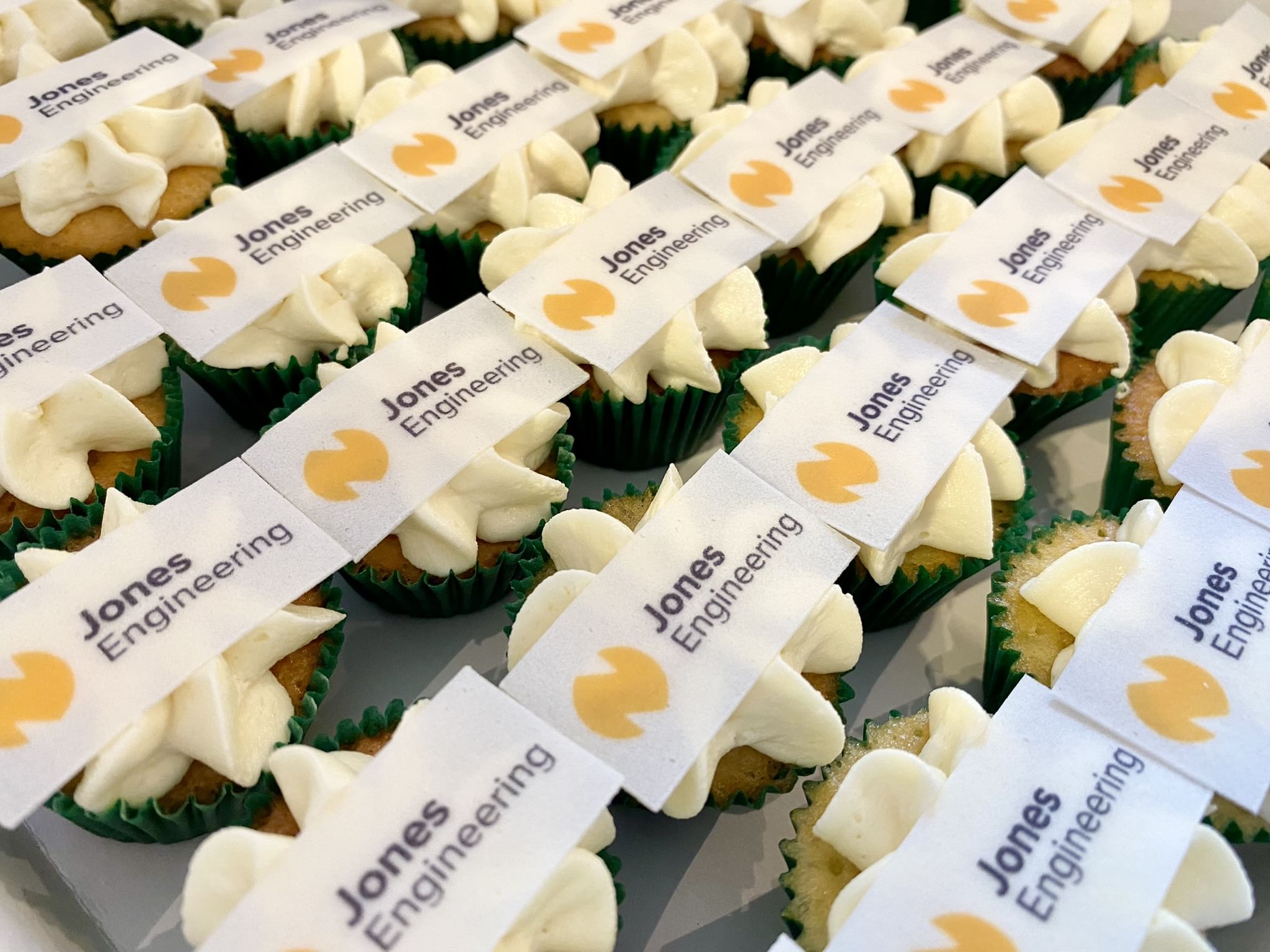 A close-up view of multiple mini vanilla cupcakes decorated with white frosting, each topped with a small rectangular edible sheet displaying the text 'Jones Engineering' and a yellow logo. Perfect for corporate events, the cupcakes are arranged in rows on a flat surface.